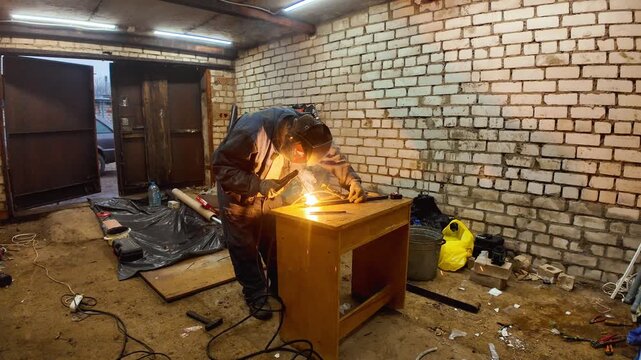 Industrial worker wearing a protective mask welding metal parts in a messy garage, with bright sparks flying from the electrode during the fabrication process in the workshop