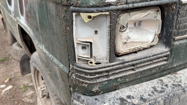 Rusted dark green minibus broken headlight, parked in gravel lot during spring, flaking paint and moss covering corroded body