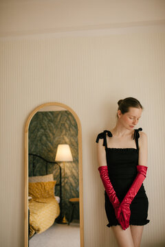 Elegant freckled woman in black dress and red gloves posing in vintage interior with soft lamp light.