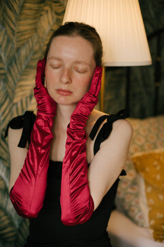 Elegant freckled woman in black dress and red gloves posing in vintage interior with soft lamp light.