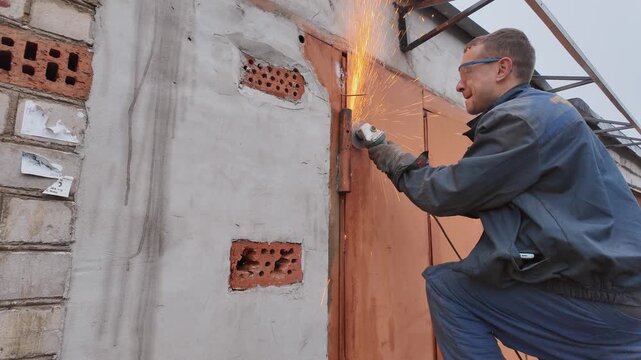 Professional male worker in protective glasses and gloves using an angle grinder to cut through a rusty metal door hinge, creating a shower of bright orange sparks against the brick wall