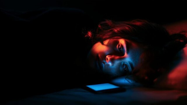Young woman lying in dark bedroom with glowing smartphone screen illuminating her face in blue and red light. Insomnia, social media addiction, dopamine, digital dependency and sleepless night concept
