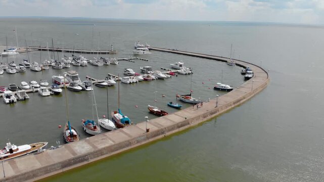 High angle aerial shot capturing numerous sailboats and motorboats moored along a curved concrete pier in a picturesque marina. The calm water reflects the cloudy sky, and a few people can be seen wal