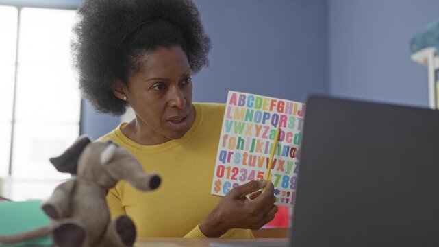 Woman teacher points with pencil to alphabet chart while holding paper beside laptop in building; learning warmth.