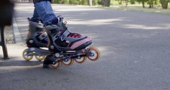 Boy sits on bench wiggling inline rollerblades above park path. Child lifts feet from pavement testing wheel movement during playful skating break after ride