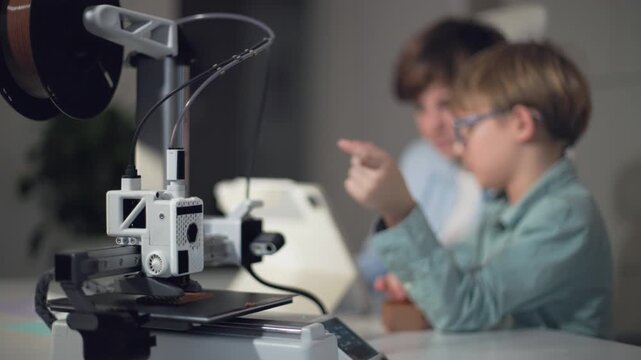 Schoolchildren at a table with a working 3D printer, observing and operating it