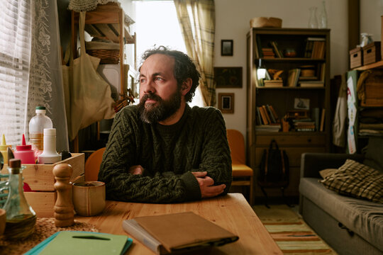 Middle aged Caucasian man sitting at table, looking through window, reflecting on financial hardship and uncertainty. Useful for poverty, unemployment, debt, recession campaigns