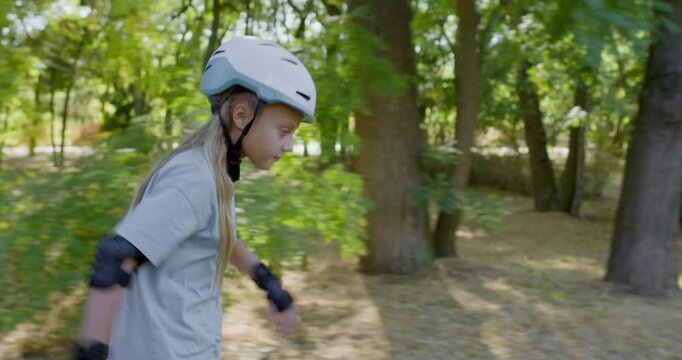 Girl skates along green park path wearing helmet and protective pads. Child leans forward keeping arms ready for balance during smooth inline rollerblade ride