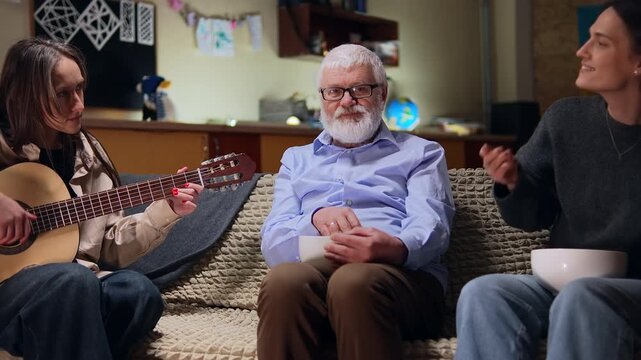 Grandpa with popcorn is surrounded by his granddaughters who sing on guitar