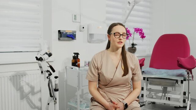 Woman gynecologist in scrub conducts professional consultation in clinic office slow motion. Doctor talks as sits near colposcope and gyno chair