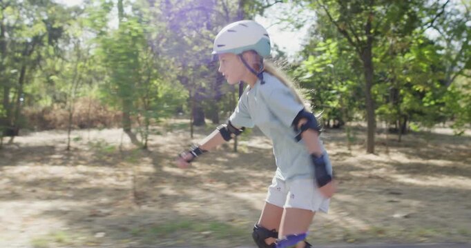 Girl skates through park path wearing helmet and protective pads. Child bends arms for balance gliding forward on rollerblades along paved walkway confidently