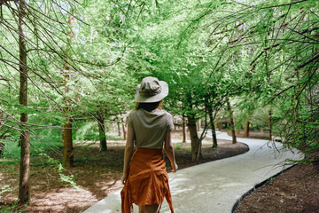 Fototapeta premium Woman walking on a curved path through green trees in a park, wearing a hat and orange skirt, back view exploring nature and summer outdoors with peaceful leafy canopy.
