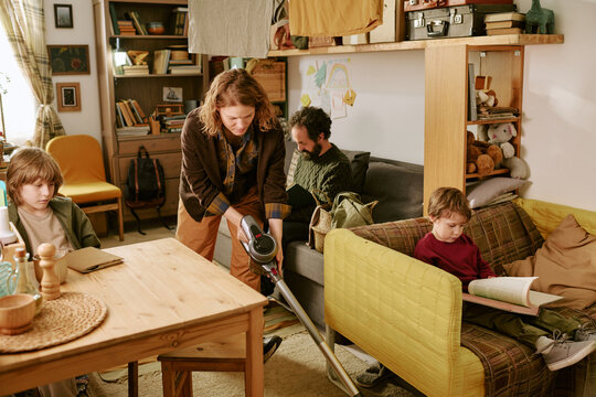 Middle aged Caucasian woman vacuuming living room while family reading and working at home, showing low income household routine. Useful for poverty, budgeting, family finance themes