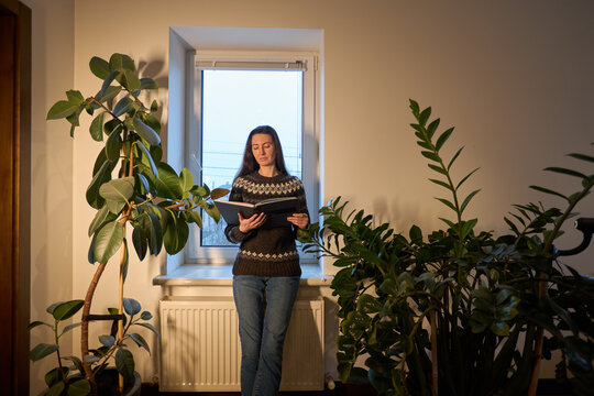 Woman reading a book indoors surrounded by lush green houseplants near a window