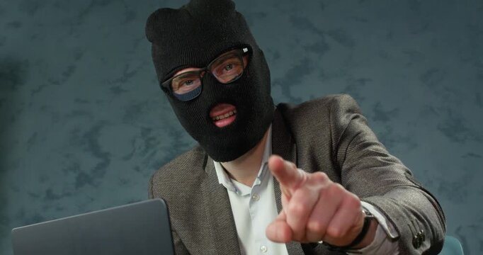 Masked scammer smiles while pointing forward near laptop during online fraud. Man directs index finger toward victim as using confident gesture to threaten target