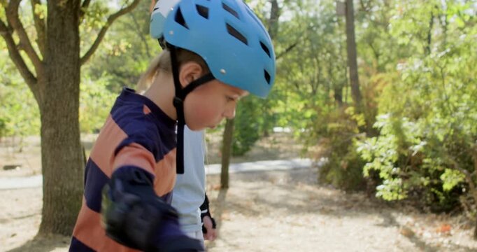 Brother and sister skate together on park path while holding hands. Children follow route near training cones keeping balance steady during shared rollerblade lesson