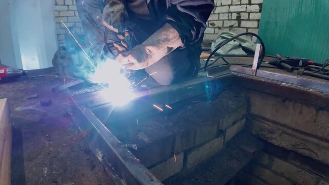 Professional welder joining metal parts with an electrode, creating bright sparks and smoke. Manual labor in a workshop during the fabrication of a steel structure for a basement hatch