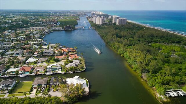 Aerial view of the Intracoastal Waterway with a boat cruising past luxury waterfront mansions and green vegetation near the Atlantic coast in Boca Raton, Florida, United States.