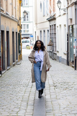 Confident young woman walking down historic cobblestone street in European city wearing trendy trench coat and casual outfit © Bjorn B
