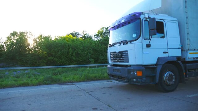 Close up of truck with cargo trailer driving on highway and transporting goods. Lorry speeding through countryside at sunset time. Cargo transportation concept. Beautiful background. Slow mo Side view