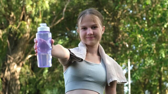 Portrait of smiling girl holding water bottle in sunny park. Concept of happiness and wellness through sport.