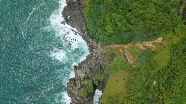 Aerial view of Bukit Pengilon featuring steep rocky cliffs and turquoise waves crashing against the shore in Kecamatan Tepus, Daerah Istimewa Yogya, Indonesia.
