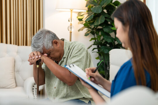 Senior Asian man looking stressed and depressed while talking to female nurse at home. Professional caregiver taking notes on clipboard, providing mental health support for elderly patient