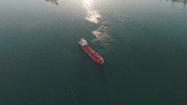 Aerial view of a red cargo ship navigating the calm blue waters near the industrial port of Augusta with sunlight reflecting on the surface in Syracuse, Sicily, Italy.