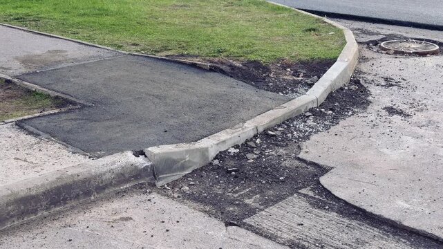 Damaged asphalt curb corner with aggregate, scattered loose asphalt and gravel along cracked pavement next to grassy verge