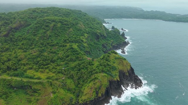 Aerial view of Bukit Pengilon featuring lush green hills and steep cliffs meeting the crashing waves of the ocean in Kecamatan Tepus, Daerah Istimewa Yogya, Indonesia.