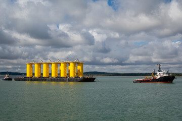 Offshore Wind Turbine Yellow Monopile Foundations on Transport Barge Towed by Tugboat at Sea....
