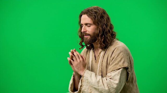 Man portraying Jesus Christ praying with closed eyes and clasped hands wearing a traditional rustic beige robe against a green screen background showing deep christian faith and religious devotion