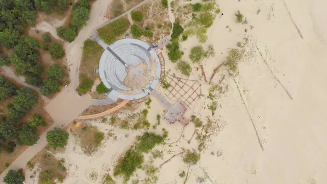 Drone footage capturing a bird's eye view of a modern, circular architectural structure, likely an observation platform or monument, situated in a natural landscape featuring sand dunes and green tree