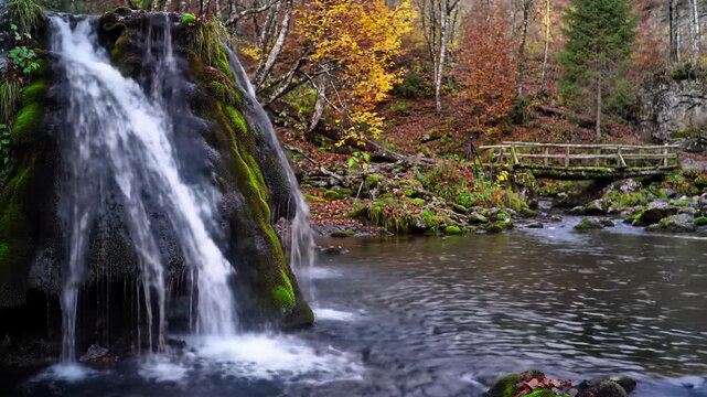 hyperrealistic wide video of bigar waterfall: bell-shaped emerald mossy rocks, crystal water cascading into a black pool, fiery autumn trees, rustic bridge, tranquil ethereal nature