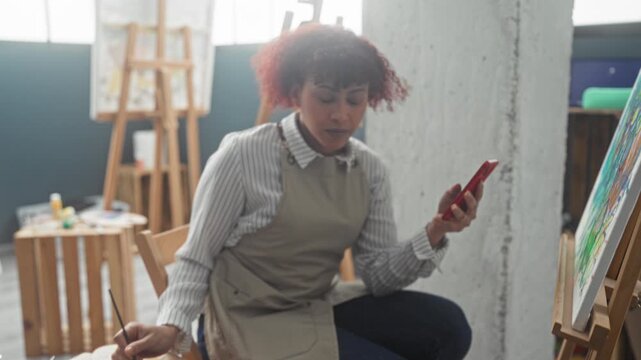 Woman painting colorful canvas with brush while holding smartphone and checking reference images in art studio; creative focus calm.