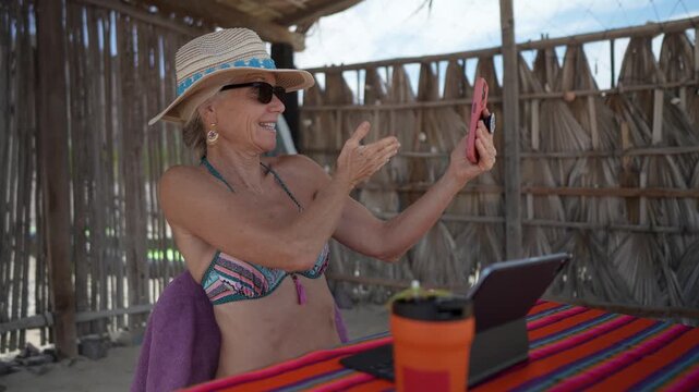 Smiling woman making video call on phone at beachside table in a tropical vacation spot.