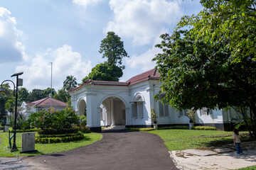 Obraz premium The historic Melchior Building, built in 1884, stands within Bogor Botanical Gardens featuring white colonial architecture, arched entryways, and red-tiled roofing surrounded by tropical greenery.
