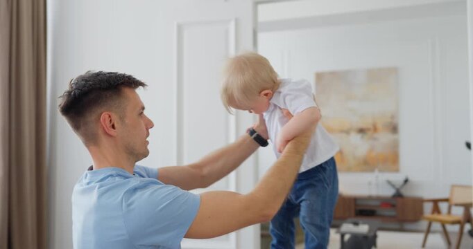 Young father lifting his smiling baby in a bright modern living room, sharing a tender family bonding moment at home near large windows