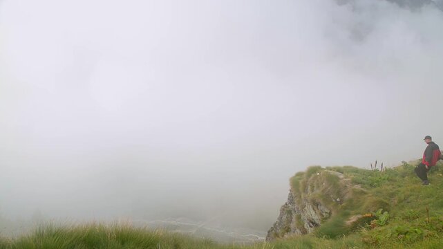 Alpine hiker standing on mountain cliff above dense fog in bad weather landscape, cinematic adventure trekking scene with mist, alpine meadows and rocky slope