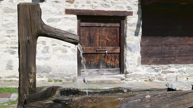 Fresh cold spring water flowing from traditional wooden mountain fountain with alpine chalet background on sunny summer day, rustic log fountain, clear drinking water, mountain countryside scene