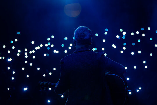 Silhouette of a guitarist playing acoustic guitar on stage during a live concert in front of an audience with phone lights
