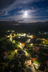 Obraz premium Aerial night view of Agioi Douloi village in Corfu, Greece, showing illuminated houses and streets nestled in the mountains under a dramatic cloudy sky and full moon.