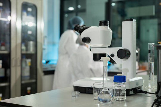 Close-up of a stereo microscope on lab bench with glassware, while masked scientists in white cleanroom suits work in a professional research facility