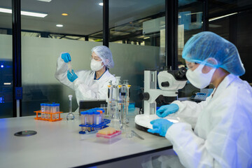 Naklejka premium Two lab technicians wearing white coats, masks, and gloves analyze specimens beside a microscope in a professional biomedical research facility