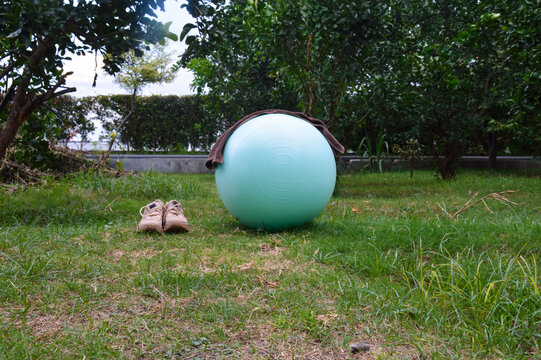 A teal exercise stability ball sits on a lush green lawn with a white towel draped on top, accompanied by brown cream athletic sneakers in an open public park.