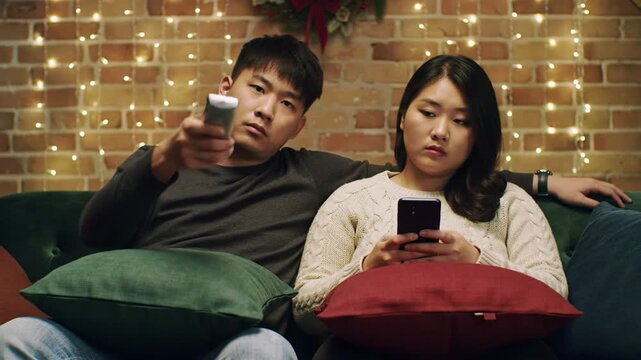 A static medium video of a young asian couple, bored and disengaged on a plush sofa in a dimly lit living room with glowing fairy lights and a christmas wreath. Mundane domestic life concept.