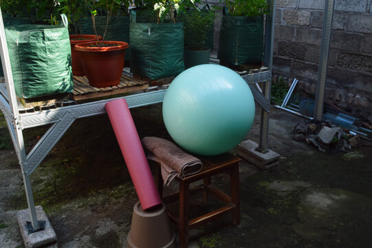 A high-angle view looking down from the front-left onto a teal gym ball, a small white towel, and a rolled pink mat arranged near a steel garden structure with shade netting.