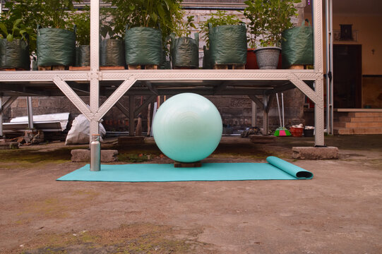 A straight eye-level front view shows an unrolled blue exercise mat holding a metallic drinking bottle alongside a large teal stability ball by a shaded garden structure.
