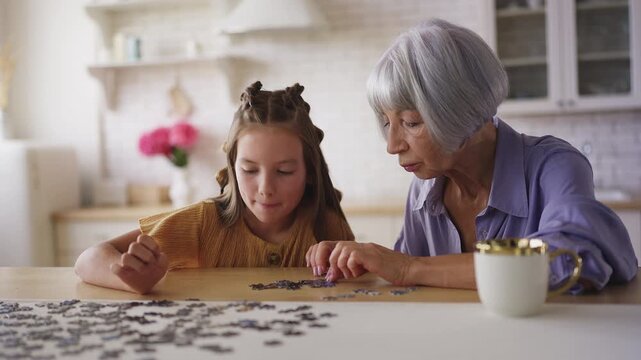 Adorable girl and her granny solving jigsaw puzzle together, family pastime