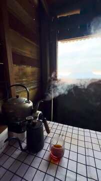 A kettle of boiling water sits on a table with a pitcher of hot water and a cup of black coffee. A wooden house with a view of the Doi Luang Chiang Dao mountain peak in Chiang Mai, Thailand.